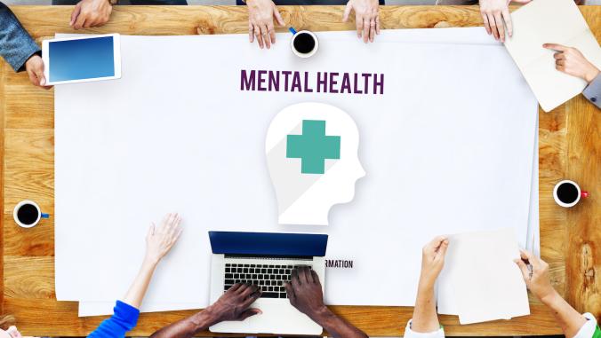 People (with only forehands and hands showing) sitting around a work table. The top poster (on a stack of posters) on the table displays the words "mental health."